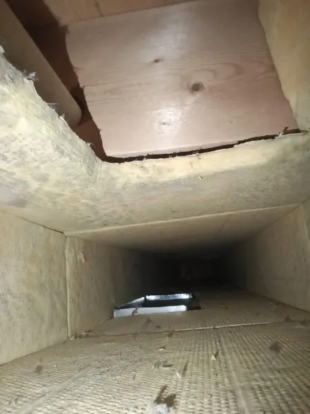 Inside a panned wood-joist return duct showing dust, debris, and a gapped wood panel before cleaning.