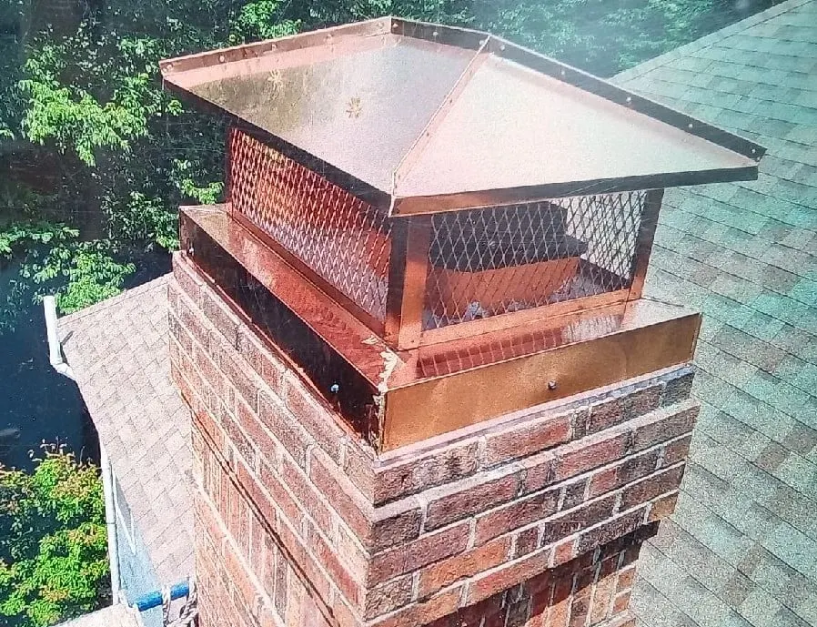 Copper chimney cap with a hip-and-ridge roof and full skirt covering the upper brick courses of the chimney.