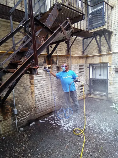 Masked Armstrong technician cleaning a dryer vent at a brick apartment building, with removed lint dusting the ground.