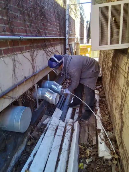 Technician snaking a cleaning rod through vent ducting in a narrow alley between brick buildings.