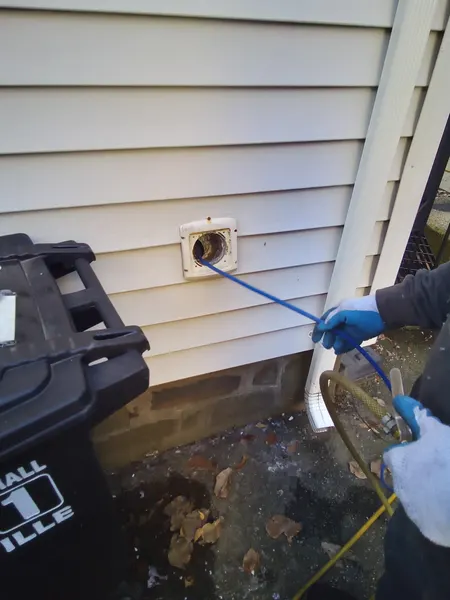 Technician feeding a flexible cleaning rod into an exterior dryer vent near the home's foundation.