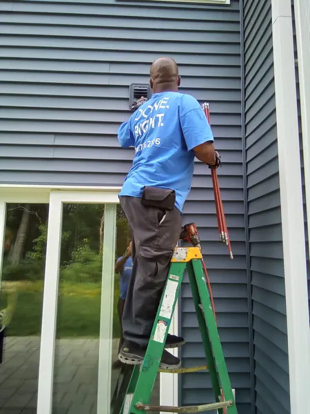 Technician on a stepladder using flexible rotary rods to brush out a wall dryer vent from outside.