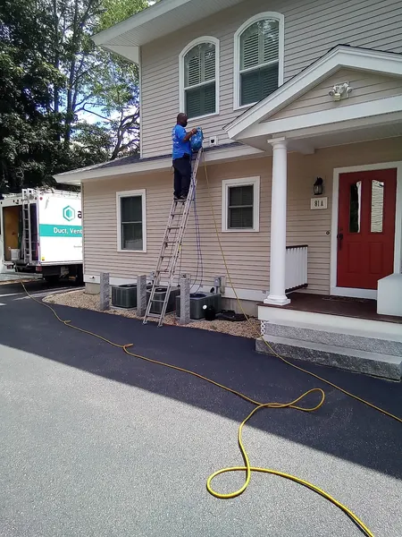 Technician on an extension ladder reaching a second-story dryer vent, with the air line run from the Armstrong truck.